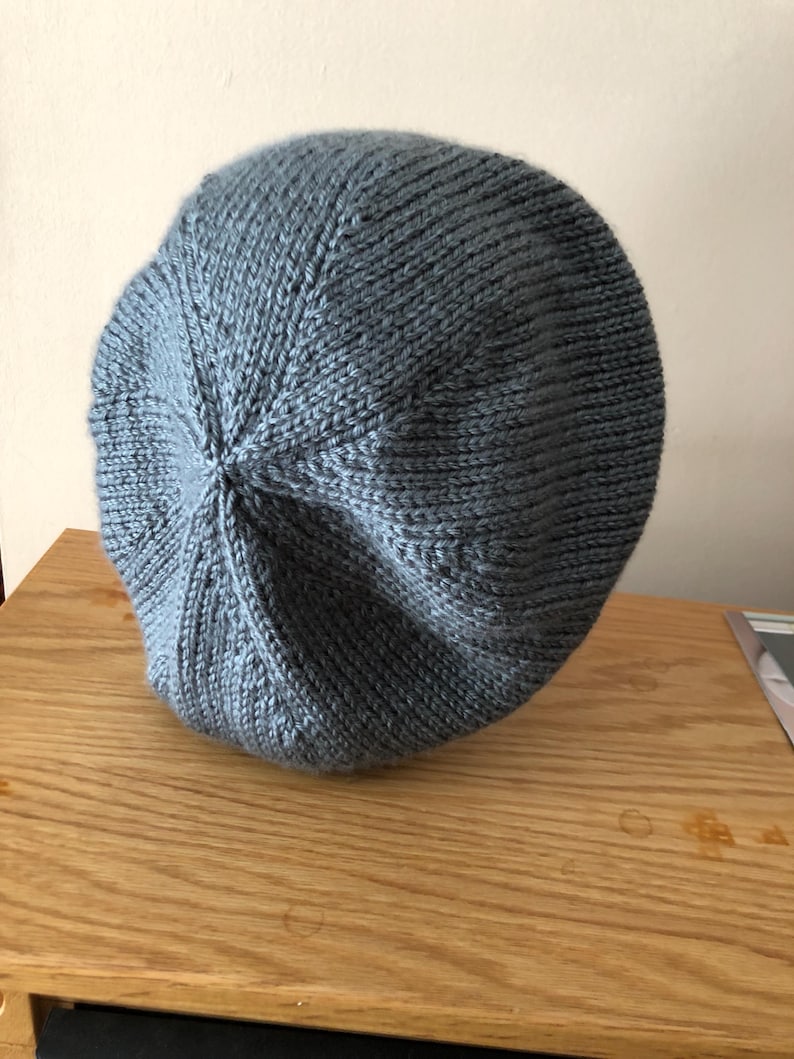 May include: A knitted, light blue beret hat. The hat is made of a soft, textured yarn and has a rounded crown. The beret is resting on a wooden surface.