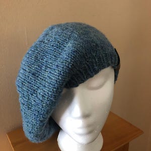 May include: A blue knit beret with a textured appearance. The hat is displayed on a white mannequin head. A small, dark brown leather accent is visible on the side of the beret. The beret is made of a soft, possibly wool, material.