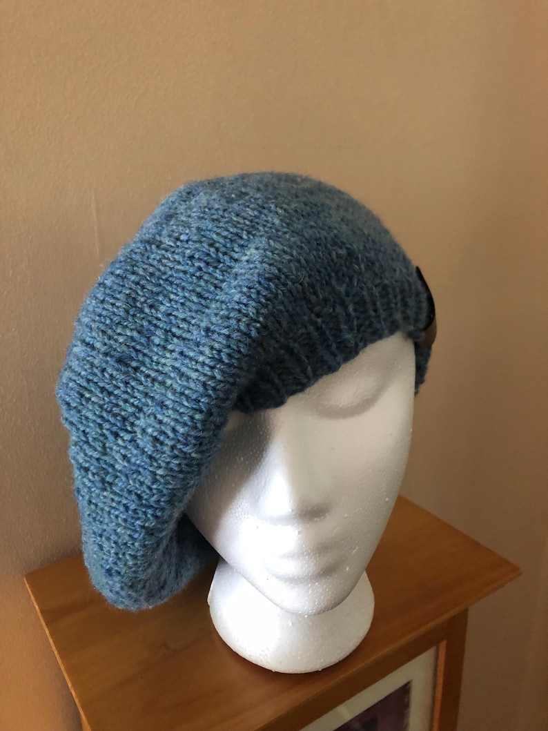 May include: A blue knit beret with a textured appearance. The hat is displayed on a white mannequin head. A small, dark brown leather accent is visible on the side of the beret. The beret is made of a soft, possibly wool, material.