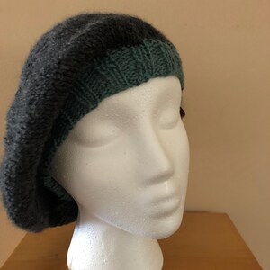 May include: A dark gray knitted beret with a forest green brim. The beret is displayed on a white mannequin head. The hat has a soft, textured appearance, suggesting a warm and comfortable accessory. This hat is perfect for adding a touch of style to any outfit.