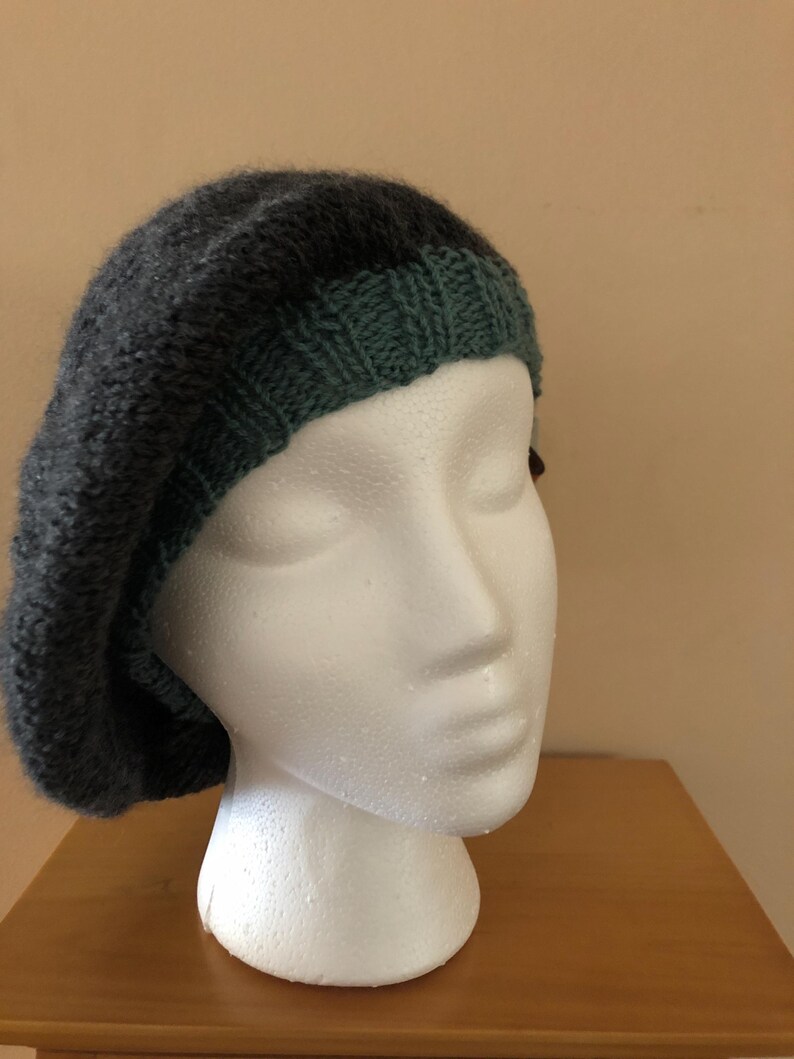 May include: A dark gray knitted beret with a forest green brim. The beret is displayed on a white mannequin head. The hat has a soft, textured appearance, suggesting a warm and comfortable accessory. This hat is perfect for adding a touch of style to any outfit.