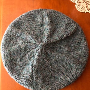 May include: A hand-knitted beret in shades of blue, green, and gray. The hat is round with a central swirl detail, showcasing a textured knit pattern. The beret is made of soft yarn, perfect for cool weather.