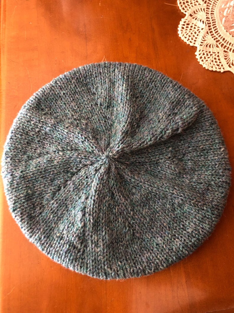 May include: A hand-knitted beret in shades of blue, green, and gray. The hat is round with a central swirl detail, showcasing a textured knit pattern. The beret is made of soft yarn, perfect for cool weather.