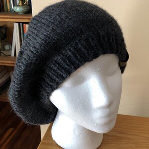 May include: A dark gray knitted beret hat. The hat is displayed on a white mannequin head. The beret has a ribbed brim and a soft, textured appearance. The hat is made of a thick, warm yarn, perfect for cold weather.