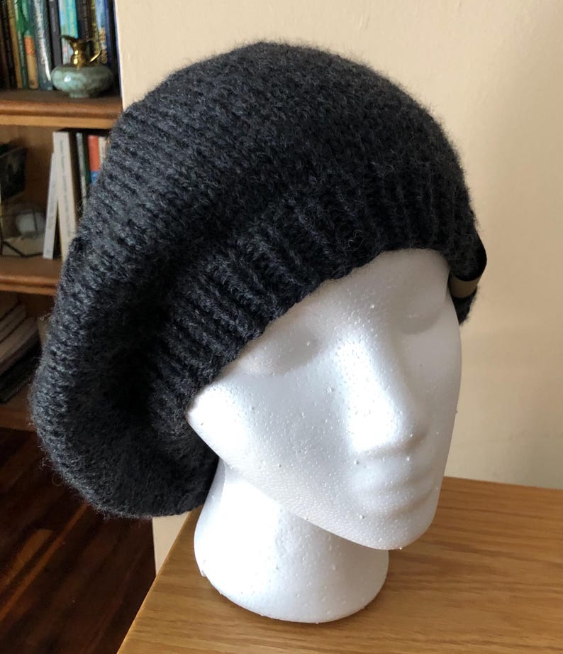 May include: A dark gray knitted beret hat. The hat is displayed on a white mannequin head. The beret has a ribbed brim and a soft, textured appearance. The hat is made of a thick, warm yarn, perfect for cold weather.
