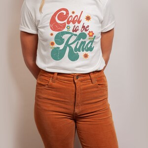 Pode incluir: Uma t-shirt branca de manga curta com o texto Cool to be Kind numa fonte vermelha e verde desgastada, decorada com pequenas ilustra&ccedil;&otilde;es de flores coloridas. A t-shirt &eacute; combinada com cal&ccedil;as de veludo cotel&ecirc; cor de laranja com fecho.