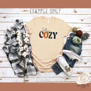 Peut inclure: Un t-shirt beige avec le texte "Stay Cozy" en lettres stylis&eacute;es color&eacute;es. Le t-shirt est pos&eacute; &agrave; plat sur une surface en bois blanc avec une chemise &agrave; carreaux, des boules de coton, un jean et des baskets.
