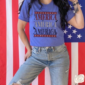 May include: A blue t-shirt with the word "AMERICA" repeated four times in a vintage style font. The shirt is decorated with red, white, and blue stars and stripes. The shirt is being worn by a person standing in front of an American flag.
