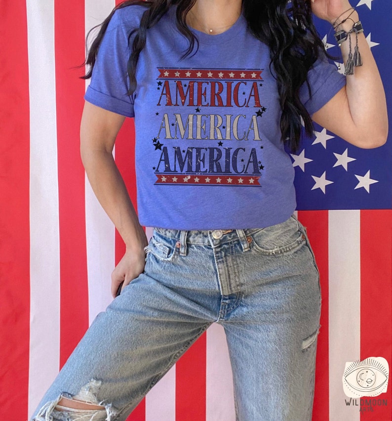 May include: A blue t-shirt with the word "AMERICA" repeated four times in a vintage style font. The shirt is decorated with red, white, and blue stars and stripes. The shirt is being worn by a person standing in front of an American flag.