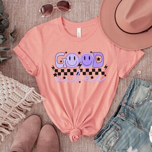 May include: A pink t-shirt with the text "Good Vibes" in purple with a checkered pattern and three purple smiley faces.