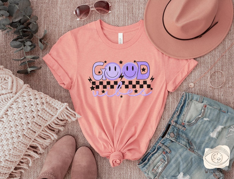 May include: A pink t-shirt with the text "Good Vibes" in purple with a checkered pattern and three purple smiley faces.