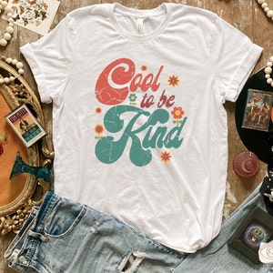Pode incluir: Uma t-shirt branca de manga curta com o texto Cool to be Kind numa fonte de estilo retro desgastada. A palavra Cool &eacute; vermelha e a palavra Kind &eacute; azul-petr&oacute;leo, acentuada por pequenas ilustra&ccedil;&otilde;es de flores coloridas em laranja, amarelo e vermelho.