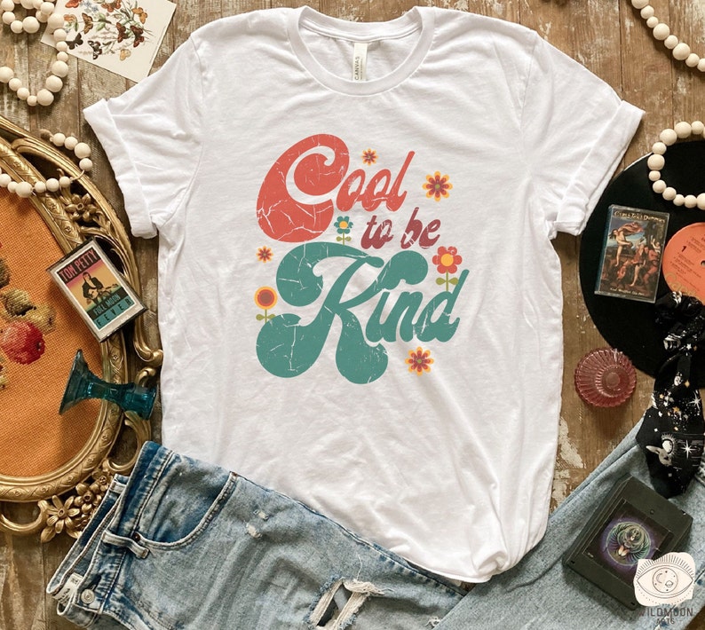 Pode incluir: Uma t-shirt branca de manga curta com o texto Cool to be Kind numa fonte de estilo retro desgastada. A palavra Cool &eacute; vermelha e a palavra Kind &eacute; azul-petr&oacute;leo, acentuada por pequenas ilustra&ccedil;&otilde;es de flores coloridas em laranja, amarelo e vermelho.