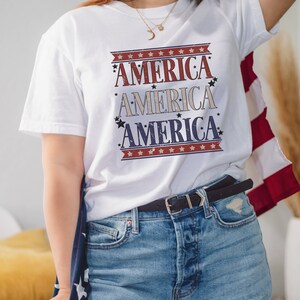 May include: A white t-shirt with a red, white, and blue graphic that says "America" three times. The shirt is being worn by a person with long brown hair and a black hat. The person is holding a red, white, and blue American flag.