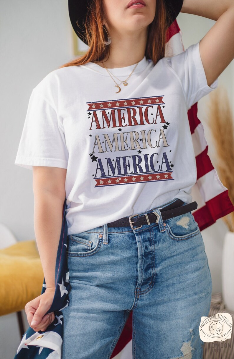 May include: A white t-shirt with a red, white, and blue graphic that says "America" three times. The shirt is being worn by a person with long brown hair and a black hat. The person is holding a red, white, and blue American flag.