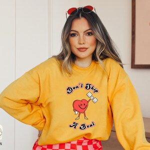 K&ouml;nnte beinhalten: Gelber Pullover mit einem roten Herz-Motiv, das eine Hantel hebt. Der Schriftzug lautet "Don't Skip A Beat".