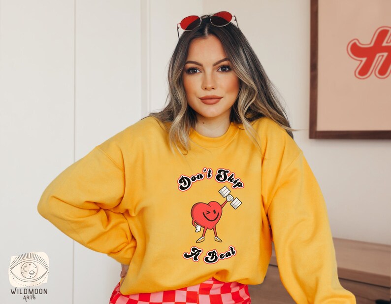K&ouml;nnte beinhalten: Gelber Pullover mit einem roten Herz-Motiv, das eine Hantel hebt. Der Schriftzug lautet "Don't Skip A Beat".