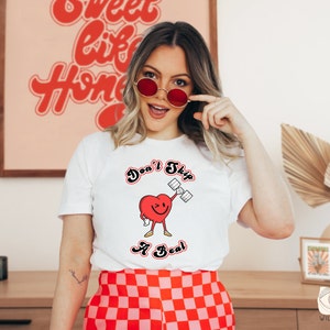 K&ouml;nnte beinhalten: Wei&szlig;es T-Shirt mit einem roten Herz-Motiv, das eine Hantel hebt. Der Schriftzug lautet "Don't Skip a Beat".