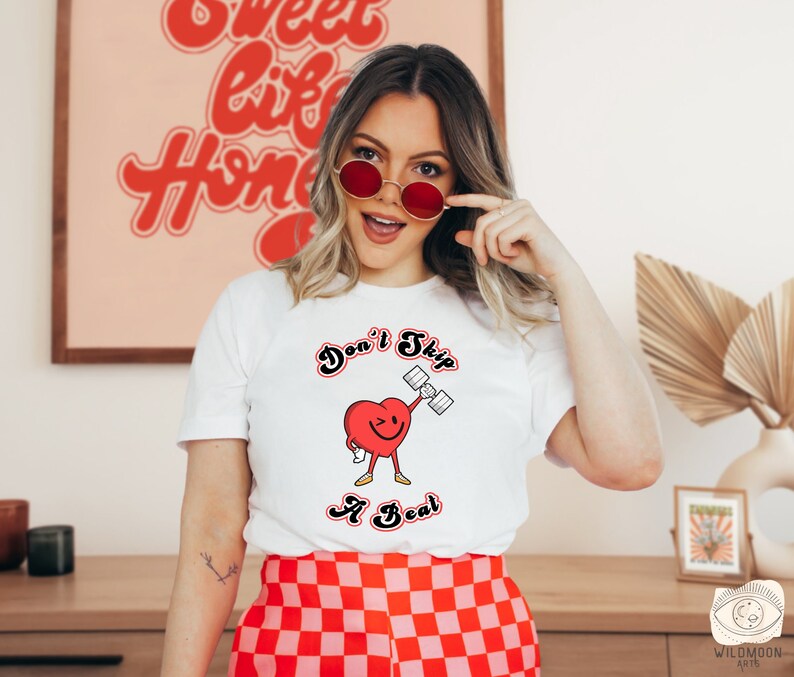 K&ouml;nnte beinhalten: Wei&szlig;es T-Shirt mit einem roten Herz-Motiv, das eine Hantel hebt. Der Schriftzug lautet "Don't Skip a Beat".