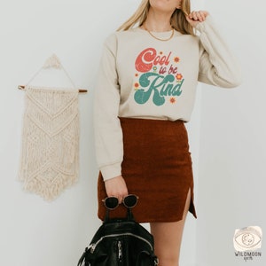Pode incluir: Uma camisola bege de manga comprida com o texto Cool to be Kind em letras vermelhas e verdes de estilo retro, decorada com pequenos gr&aacute;ficos de flores coloridas. A camisola &eacute; combinada com uma saia de veludo cotel&ecirc; castanha, uma mochila preta e &oacute;culos.