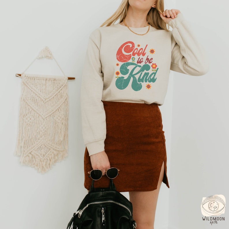 Pode incluir: Uma camisola bege de manga comprida com o texto Cool to be Kind em letras vermelhas e verdes de estilo retro, decorada com pequenos gr&aacute;ficos de flores coloridas. A camisola &eacute; combinada com uma saia de veludo cotel&ecirc; castanha, uma mochila preta e &oacute;culos.