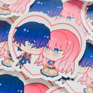 May include: A pile of heart-shaped stickers with anime-style illustrations. Each sticker depicts a couple with blue and pink hair, blue eyes, and detailed outfits. The stickers are outlined with a pink heart.