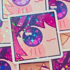 Pode incluir: Autocolantes quadrados com um olho de estilo anime estilizado com detalhes em rosa, roxo e azul. Os autocolantes têm um acabamento holográfico e brilhante com brilhos em forma de estrela. O fundo é de cor pêssego claro.