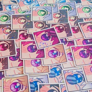 Pode incluir: Uma cole&ccedil;&atilde;o de autocolantes quadrados com olhos estilizados em estilo anime em v&aacute;rias cores. Os autocolantes t&ecirc;m um acabamento hologr&aacute;fico. Os desenhos dos olhos incluem azul, verde, roxo e rosa. Os autocolantes est&atilde;o espalhados.