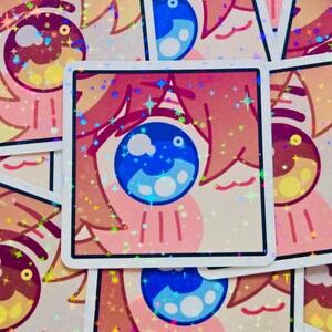 Pode incluir: Autocolantes quadrados com um desenho de olho de desenho animado. O olho tem uma &iacute;ris azul e detalhes em rosa e p&ecirc;ssego. Os autocolantes t&ecirc;m um efeito hologr&aacute;fico e brilhante com brilhos em forma de estrela. Os autocolantes est&atilde;o empilhados.