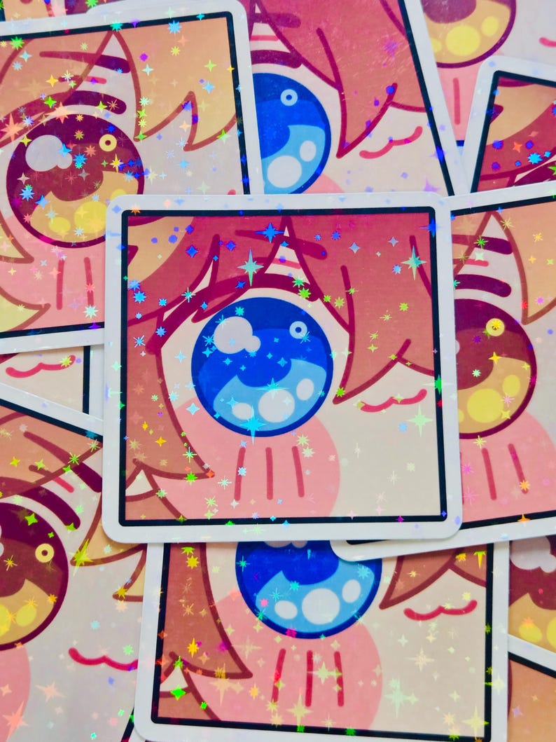 Pode incluir: Autocolantes quadrados com um desenho de olho de desenho animado. O olho tem uma &iacute;ris azul e detalhes em rosa e p&ecirc;ssego. Os autocolantes t&ecirc;m um efeito hologr&aacute;fico e brilhante com brilhos em forma de estrela. Os autocolantes est&atilde;o empilhados.