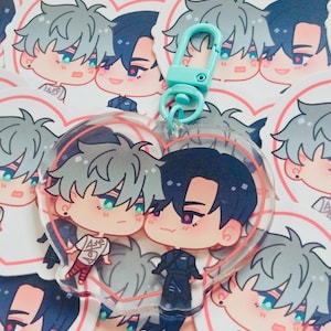 May include: A clear acrylic heart-shaped keychain featuring two cartoon characters with gray and black hair, and teal eyes. The keychain has a teal clasp. The background is covered with heart-shaped stickers with the same cartoon characters.