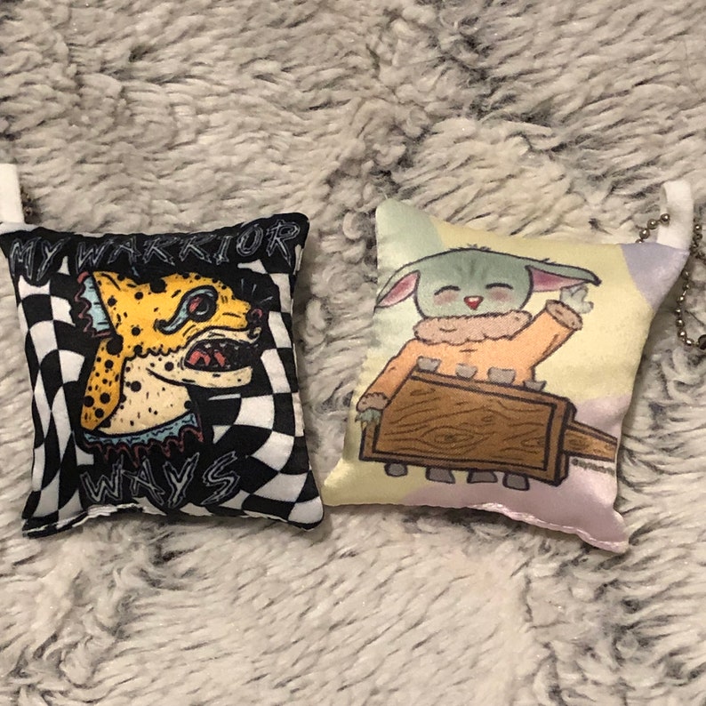 May include: Two small, colorful, plush pillows with printed designs. One pillow features a black and white checkered background with a yellow and blue cartoon animal with the text "My Warrior Ways". The other pillow features a light green cartoon character with a light brown background and a light blue, pink, and yellow background.