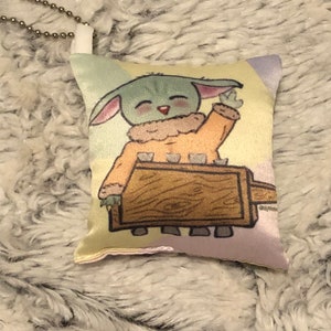May include: A small, plush pillow with a cartoon image of a green alien character with large ears, wearing a brown and orange outfit, holding a wooden stick. The pillow has a light pink, yellow, and green background.