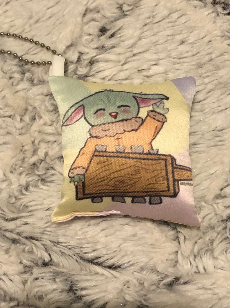 May include: A small, plush pillow with a cartoon image of a green alien character with large ears, wearing a brown and orange outfit, holding a wooden stick. The pillow has a light pink, yellow, and green background.