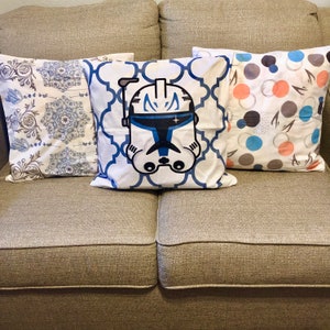May include: Three decorative throw pillows on a brown couch. The center pillow features a white background with a blue and white patterned border and a cartoon image of a blue and white Star Wars clone trooper helmet. The pillow on the left has a white background with a blue and white floral pattern. The pillow on the right has a white background with a blue, gray, and peach polka dot pattern.