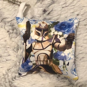 May include: A white pillow with a blue floral pattern and a printed image of a Star Wars clone trooper. The trooper is wearing a white and brown helmet and armor. The pillow is approximately 4 inches by 5 inches.