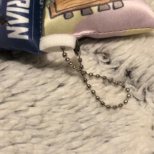 May include: A small, white, plush pillow with a colorful cartoon design on one side and the words "The Magic of Lorian" on the other side. The pillow has a white zipper and a silver chain attached to it.