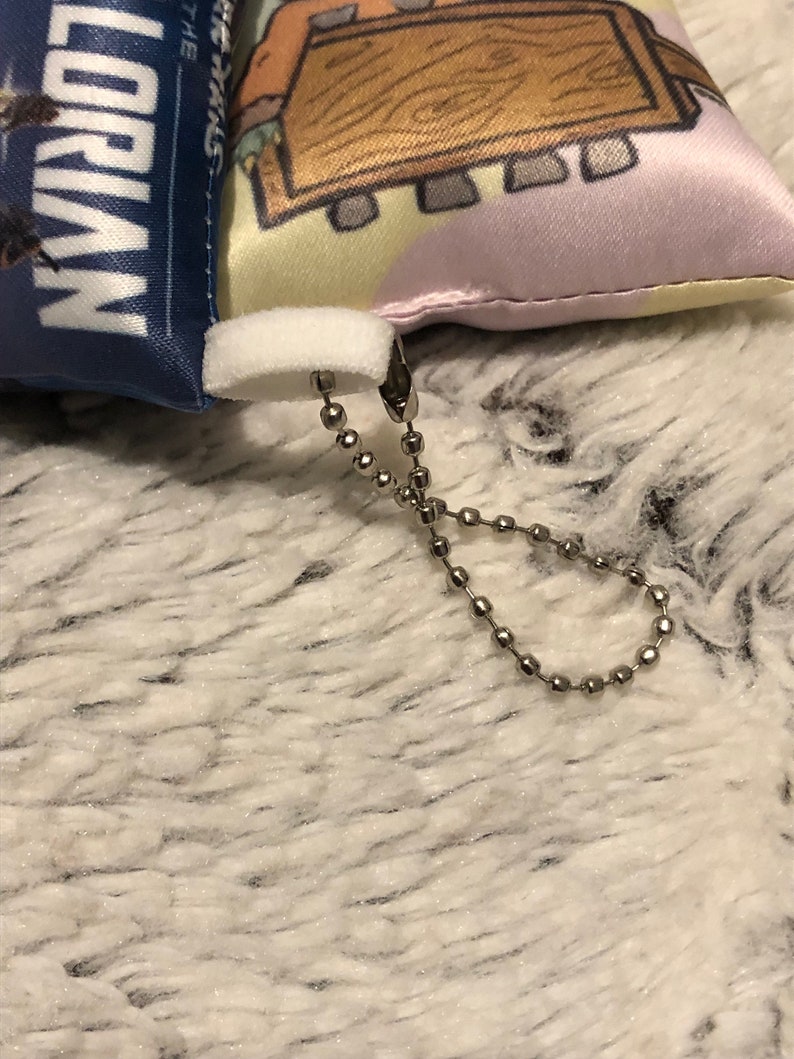 May include: A small, white, plush pillow with a colorful cartoon design on one side and the words "The Magic of Lorian" on the other side. The pillow has a white zipper and a silver chain attached to it.