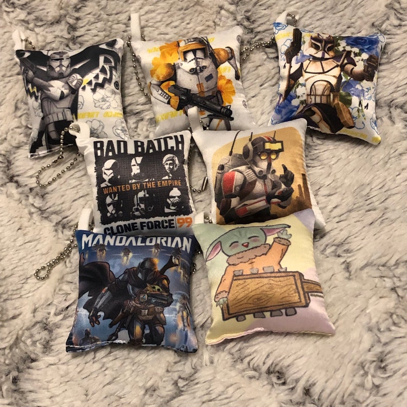 May include: Eight small, white, square pillows with different Star Wars characters printed on them. The pillows have a chain attached to them, making them keychains. The characters include:  Clone Force 99,  The Mandalorian,  Baby Yoda,  and a Stormtrooper.