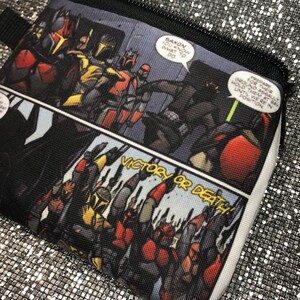 May include: A black and white comic book style zippered pouch with a red, black, and yellow graphic of characters in a battle scene. The text "VICTORY OR DEATH!" is printed in white on a black background.