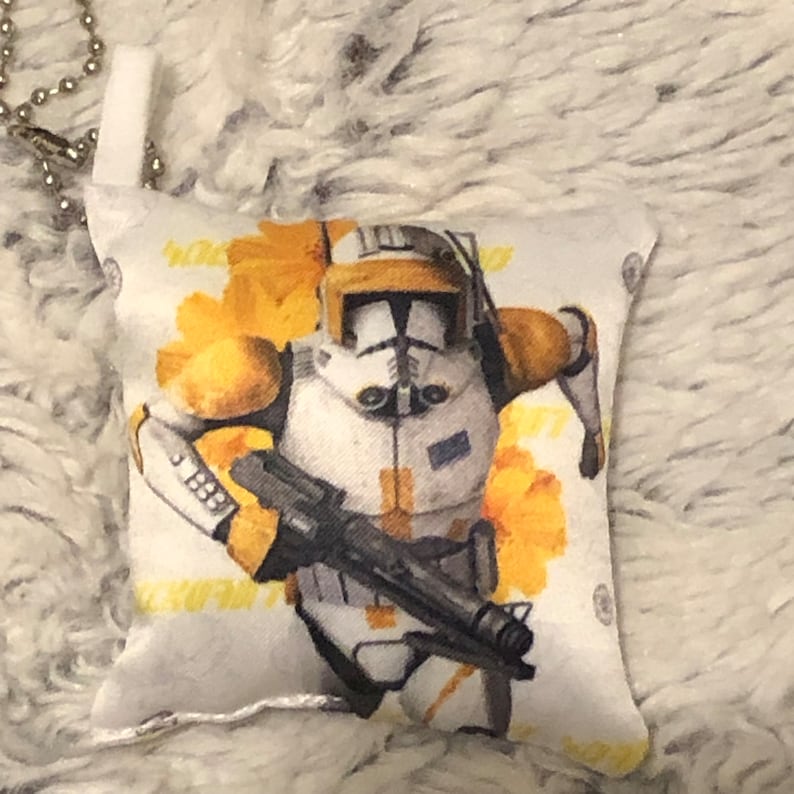 May include: A white and yellow pillow with a Star Wars character on it. The character is wearing a white helmet with a yellow visor and a white and yellow suit. The character is holding a gun in their right hand. The pillow is about 10 inches wide and 12 inches long.