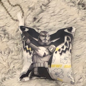 May include: A white pillow with a black and white image of a Star Wars clone trooper holding a blaster rifle. The pillow has a white background with a floral pattern and the text "Clone Wars" printed on it.