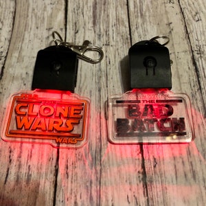 Puede incluir: Dos llaveros rojos con luz y frentes de plástico transparente. El llavero de la izquierda dice "Star Wars The Clone Wars" y el llavero de la derecha dice "Star Wars The Bad Batch".