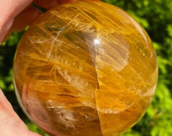 Puede incluir: Una gran esfera de citrino amarillo dorado con una superficie lisa y pulida. La esfera tiene un patrón estriado en toda su superficie, creando una textura única y hermosa.