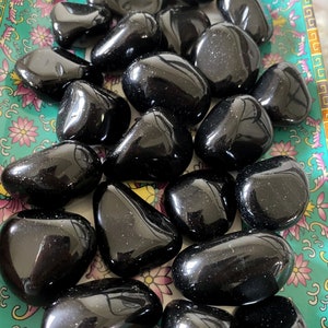 May include: A collection of black obsidian tumbled stones on a patterned surface. The stones are smooth and polished, with a glossy finish.