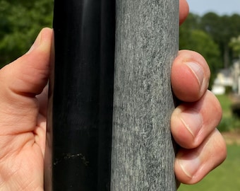 Puede incluir: Dos objetos cilíndricos de piedra, uno negro y otro gris, se sostienen en una mano. El objeto negro tiene una superficie lisa y pulida. El objeto gris tiene un aspecto texturizado y moteado. Ambos miden aproximadamente 13 cm de altura.