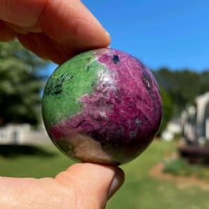 Puede incluir: Una esfera de piedra preciosa pulida, sostenida entre los dedos, que muestra una mezcla de colores. La esfera presenta una mezcla de verde, magenta intenso y motas negras, con una forma lisa y redondeada. El fondo es un cielo azul claro y hierba verde.