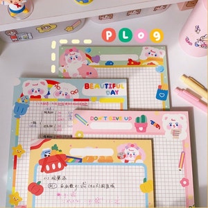 Puede incluir: Tres blocs de notas coloridos con papel cuadriculado e ilustraciones de animales de dibujos animados. El bloc de notas superior tiene un fondo azul claro con un arcoíris y el texto "PLog 9". El bloc de notas del medio tiene un fondo rosa con el texto "BEAUTIFUL DAY". El bloc de notas inferior tiene un fondo amarillo con el texto "DON'T GIVE UP".