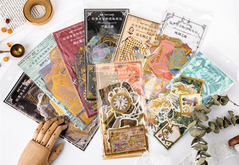 May include: A collection of decorative paper stickers in various designs, including clocks, tickets, and botanical illustrations. The stickers are packaged in clear plastic sleeves, with some featuring gold foil accents and Chinese text. The overall aesthetic is vintage and artistic.