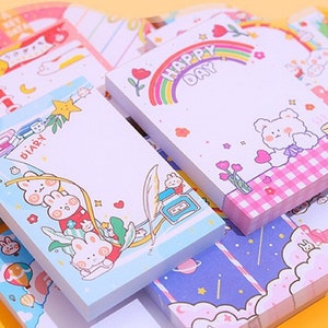 May include: A selection of colourful memo pads with different designs. Some pads feature cartoon bunnies, rainbows, and the text "HAPPY DAY" and "DIARY". The pads have a variety of pastel colours and are stacked together.